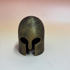 May include: A bronze-colored helmet with a rounded top and eye and mouth openings. The helmet has a textured surface and appears to be made of metal. The design includes a series of small rivets around the edges, suggesting a historical or fantasy theme.
