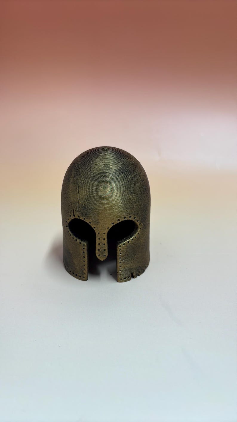 May include: A bronze-colored helmet with a rounded top and eye and mouth openings. The helmet has a textured surface and appears to be made of metal. The design includes a series of small rivets around the edges, suggesting a historical or fantasy theme.