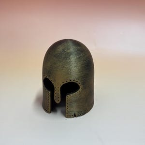 May include: A bronze-colored, miniature helmet with eye and face openings. The helmet has a rounded top and a textured surface, with small holes along the edges. It is likely a decorative item or a collectible.