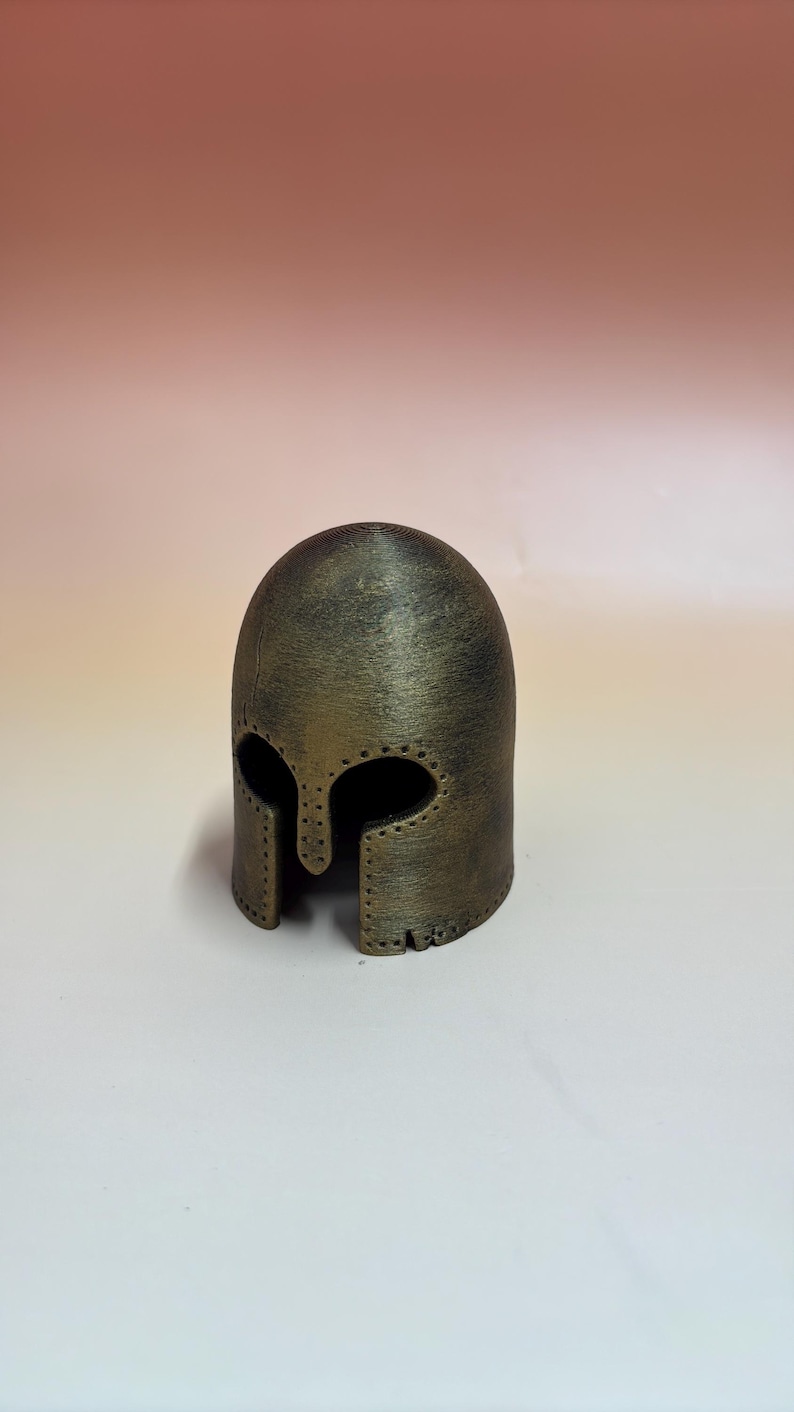 May include: A bronze-colored, miniature helmet with eye and face openings. The helmet has a rounded top and a textured surface, with small holes along the edges. It is likely a decorative item or a collectible.