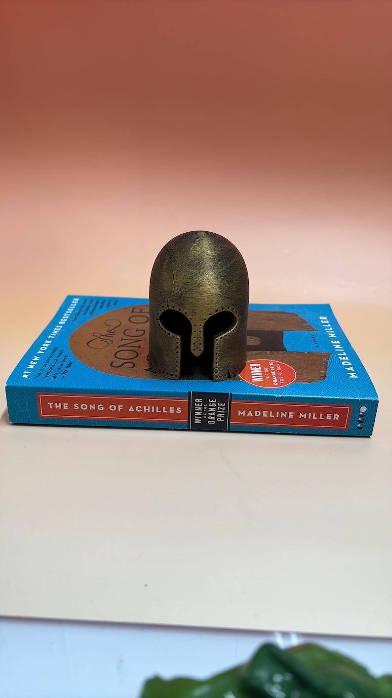 May include: A bronze-colored, miniature Spartan helmet stands on a copy of "The Song of Achilles" by Madeline Miller. The book has a blue cover with the title in large gold letters and the author's name in white.
