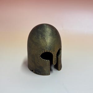 May include: A bronze-colored helmet with a rounded top and eye and mouth openings. The helmet has a textured surface and appears to be made of metal. It is a replica of a historical helmet.