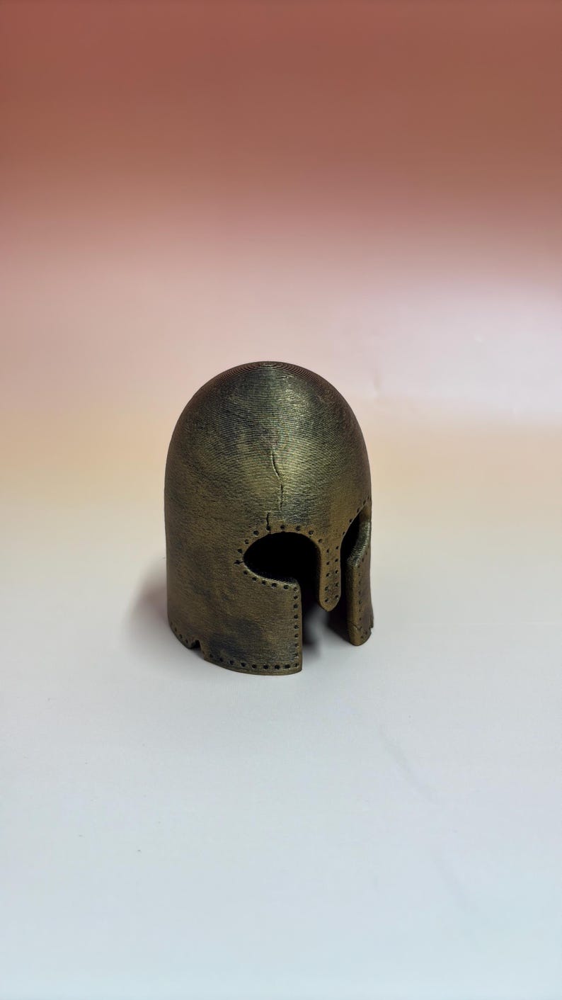 May include: A bronze-colored helmet with a rounded top and eye and mouth openings. The helmet has a textured surface and appears to be made of metal. It is a replica of a historical helmet.