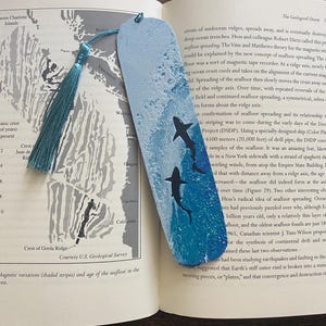 May include: Hand-painted wooden bookmark featuring a blue ocean scene with three black shark silhouettes. The bookmark has a teal tassel and is set against an open book with a map.