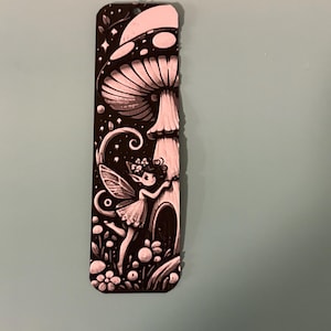 May include: Black and white decorative bookmark featuring a whimsical fairy scene. The design includes a large mushroom, stars, flowers, and a fairy with wings. The bookmark is rectangular with a rounded top.