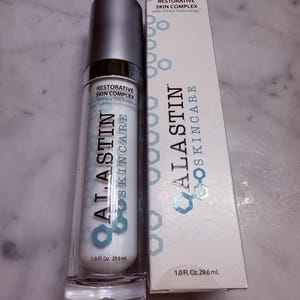 Puede incluir: Un producto para el cuidado de la piel con tapa plateada y botella blanca, etiquetado como "ALASTIN SKINCARE". El producto va acompañado de una caja blanca con la misma marca y el texto "RESTORATIVE SKIN COMPLEX". La botella contiene 29,6 ml.