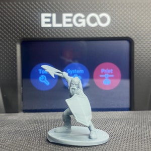 May include: A gray 3D printed miniature figure of a warrior holding a sword and shield. The figure is standing on a round base. The figure is in front of a black and gray 3D printer with a screen that says "ELEGOO" and has buttons labeled "Tool", "System", and "Print".