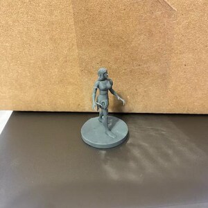 May include: A gray plastic miniature figurine of a female figure with a tattered dress and bare arms and legs. The figure is standing on a round base.