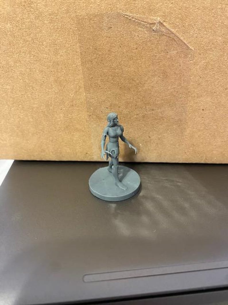 May include: A gray plastic miniature figurine of a female figure with a tattered dress and bare arms and legs. The figure is standing on a round base.