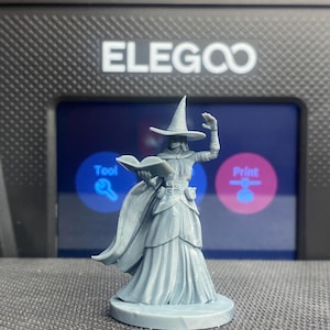 May include: A gray 3D printed miniature of a witch holding a book and wearing a pointed hat and a long robe. The miniature is standing on a round base.