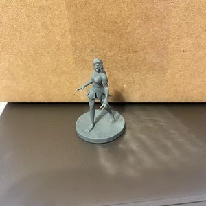 May include: A gray plastic miniature figurine of a female figure with long hair, wearing a skirt and standing on a round base.