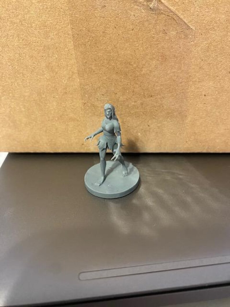 May include: A gray plastic miniature figurine of a female figure with long hair, wearing a skirt and standing on a round base.