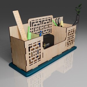 Puede incluir: Organizador de escritorio de madera con cuatro compartimentos, una regla, bol&iacute;grafos, l&aacute;pices, tijeras y una peque&ntilde;a caja negra. El organizador tiene una base azul verdosa y un patr&oacute;n geom&eacute;trico en la parte delantera.