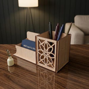 Op de afbeelding: Een bureau-organizer van lichtgekleurd hout met een vierkante pennenhouder met een lasergesneden bloemmotief aan de zijkant en een vak met een blauwe kaart waarop de tekst MGS Laser Cut Files in het wit is gedrukt.