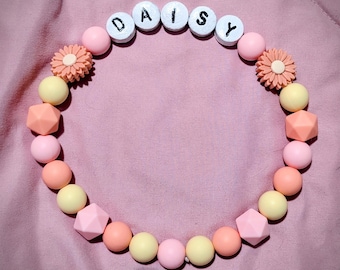以下が含まれることがあります： 白い木製のビーズで「DAISY」と書かれた円形のネックレス。 ネックレスは、ピンク、ピーチ、黄色の球体と幾何学的な形を含む、さまざまなパステルカラーのビーズと、2つの花の形をしたビーズが特徴です。