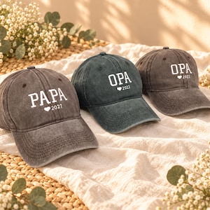Op de afbeelding: Drie baseballpetten in tinten antraciet en donkergrijs. De linker pet heeft de witte tekst PAPA met een hartje en 2027. De middelste en rechter pet hebben de witte tekst OPA met een hartje en 2021. Elke pet heeft een gebogen klep en stiksels.
