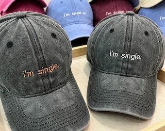 May include: Two charcoal grey baseball caps with a distressed finish. Each cap features the embroidered text 'i'm single.' on the front panel. The cap on the left has orange lettering, while the cap on the right has white lettering.