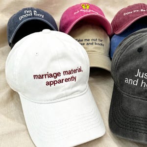 Op de afbeelding: Een collectie van zes baseballpetten in diverse kleuren, waaronder wit, donkergrijs, marineblauw, roze, beige en bordeauxrood. Elke pet is aan de voorzijde voorzien van geborduurde tekst, zoals 'marriage material, apparently' en 'I'm a good boy'.