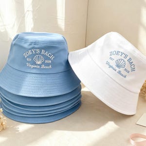May include: A stack of light blue bucket hats and one white bucket hat, each featuring white embroidered text that reads Zoey's Bach, Est 2026, Virginia Beach, and a central seashell graphic. The hats have a wide brim and a flat crown.