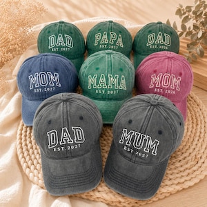 Può includere: Una collezione di nove cappellini da baseball in vari colori, tra cui verde, blu, grigio e rosa. Ogni cappellino presenta scritte bianche ricamate con titoli come DAD, PAPA, MOM, MAMA o MUM, accompagnati da un anno come EST. 2026 o 2027.