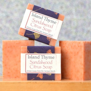 Puede incluir: Tres barras de jabón de color naranja con una etiqueta que dice "Island Thyme Sandalwood Citrus Soap Artisan Soaps from the San Juan Islands."