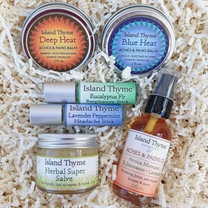 Puede incluir: Una caja de regalo llena de remedios naturales de Island Thyme. La caja contiene una variedad de productos, incluyendo un bálsamo de calor profundo, un bálsamo de calor azul, un bastón respiratorio de eucalipto y abeto, un bastón para el dolor de cabeza de lavanda y menta, una pomada súper herbal y un aceite para dolores. Todos los productos están etiquetados con el logotipo de Island Thyme y el nombre del producto.