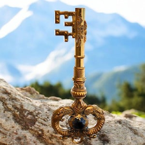 May include: A large, ornate, gold-colored key with a black gemstone in the center of the bow. The key is resting on a rock with a mountain range in the background.