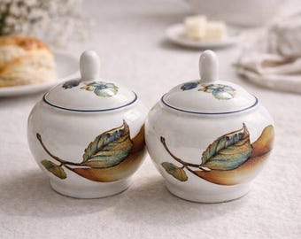 May include: Two white ceramic sugar bowls with lids. Each bowl features a leaf design in shades of brown, green, and blue. The lids have a small handle and a blue floral pattern. The bowls are on a white tablecloth.