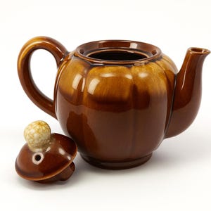 May include: A brown ceramic teapot with a matching lid. The teapot has a rounded body, a curved handle, and a spout. The lid is off to the side, revealing the opening. The teapot is a glossy brown colour.