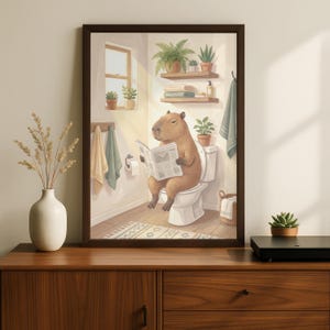 Puede incluir: Impresión artística enmarcada que muestra un carpincho sentado en un inodoro, leyendo un periódico. La escena del baño incluye plantas, toallas y una ventana con luz solar. La obra de arte está en un marco marrón.