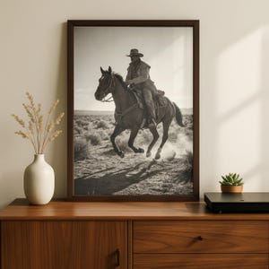 Op de afbeelding: Zwart-wit ingelijste print van een cowboy te paard in galop. De cowboy draagt een hoed en een stofjas, het paard trapt stof op. De achtergrond toont een woestijnlandschap onder een heldere hemel. De lijst is bruin.