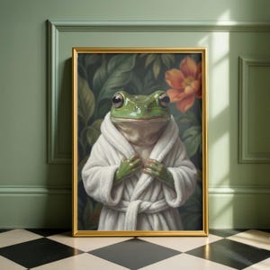 May include: A framed art print featuring a green frog wearing a white bathrobe. The frog is the focal point, set against a backdrop of lush green foliage and an orange flower. The frame is gold, adding a touch of elegance.