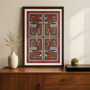May include: Framed artwork featuring a geometric design with bird-like figures in a grid pattern. The artwork uses red, brown, green, and black colors. The frame is dark brown, and the artwork is displayed on a wooden cabinet.