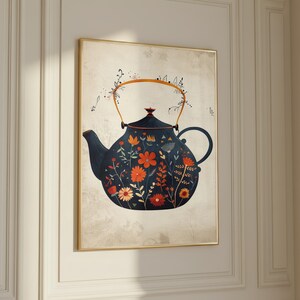 May include: Framed print of a navy blue teapot decorated with orange, yellow, and white floral patterns. The teapot has a gold handle and spout, set against a beige backdrop. The artwork is displayed on a white wall.