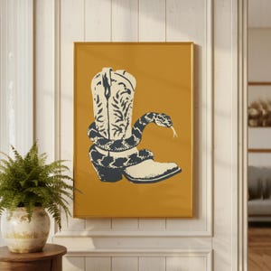 May include: A framed art print depicting a cowboy boot and a snake. The boot is off-white with dark blue floral detailing. A dark blue and off-white snake is coiled around the boot, with its tongue extended. The background is mustard yellow.