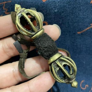May include: A vintage brass vajra, a ritual object with a central black fabric grip and leather strap. The vajra features intricate openwork designs and a pointed end. The brass has a slightly aged appearance.