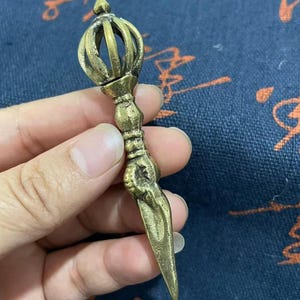 May include: A bronze-colored vajra, a ritual object with a central handle and a spiked, pointed end. The handle features intricate detailing and a spherical top. The object is held against a blue patterned background.