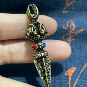 May include: A bronze-colored pendant with a detailed design. The pendant features a loop at the top, a decorative crown-like element, and a triangular, pointed bottom. It has red and blue accents and intricate patterns.