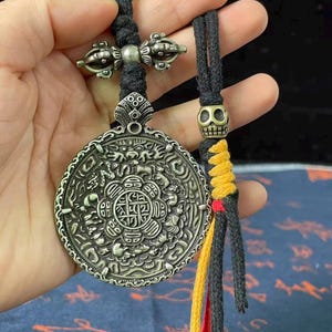 May include: A round silver-toned metal pendant featuring intricate embossed animal and geometric patterns, suspended from a black braided cord. The cord includes a brass-toned skull bead and a decorative metal vajra charm with yellow and red tassels.