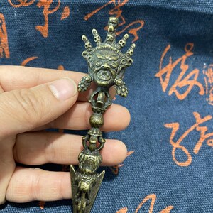 May include: A bronze-colored, ornate ritual dagger with a detailed design. The dagger features a central face with a crown-like structure above and a pointed blade. The handle has multiple decorative elements.