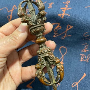 May include: A detailed bronze vajra, a ritual object, featuring intricate carvings and a central blade. The vajra has a weathered appearance, with a dark brown patina and ornate details. The design includes floral and claw-like elements.