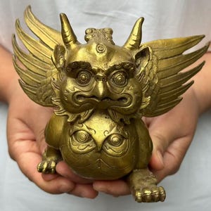 Könnte beinhalten: Eine messingfarbene Metallfigur eines Fabelwesens mit Flügeln, Hörnern und einem dritten Auge auf der Stirn. Das Wesen hat einen runden Körper, eine schnabelartige Nase und detaillierte Gesichtszüge sowie ein augenähnliches Muster auf dem Bauch.