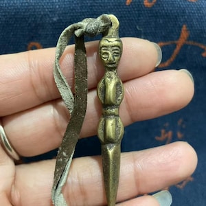 Puede incluir: Un pequeño colgante antiguo de latón con una figura humana estilizada en la parte superior y una parte inferior puntiaguda. Tiene un lazo para colgar y está unido a un cordón de cuero marrón desgastado. El colgante mide unos 5 cm de largo.