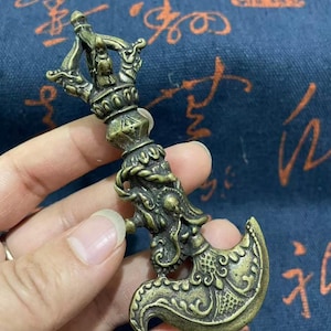 May include: A bronze-colored metal ritual object featuring a curved blade at the base and a multi-pronged top. The handle is decorated with intricate relief carvings of a dragon head and swirling patterns, showcasing a weathered, antique-style finish.