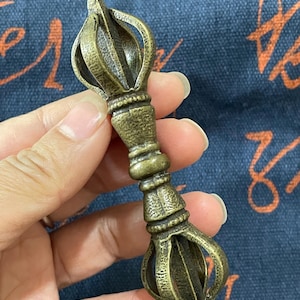 May include: A bronze-colored Vajra, a ritual object, is held in a hand. The Vajra features a central handle with decorative rings and a flared, multi-pronged top and bottom. The object has an aged, antique appearance.