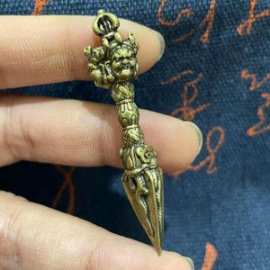 May include: A detailed bronze-colored pendant with a crown-like top and a detailed face. The pendant has a cylindrical handle with intricate carvings and a pointed, blade-like bottom. The overall design suggests a religious or ceremonial artifact.