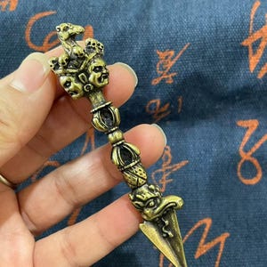 May include: A detailed bronze-colored ritual dagger with intricate carvings. The handle features multiple faces and a serpent design, while the blade is a sharp, triangular point. The dagger is held against a patterned blue background.
