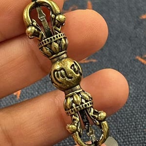 May include: A detailed close-up of a bronze-colored Vajra, a ritual object. The Vajra features intricate floral and geometric designs, with a central handle and pointed ends. The object is held in a hand, showcasing its ornate details and craftsmanship.