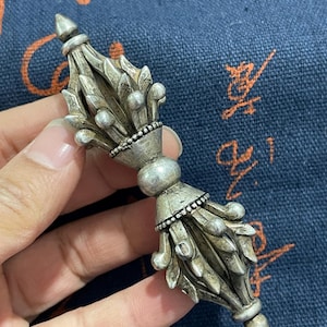 May include: A silver vajra, a ritual object with intricate detailing. The vajra has a central bulbous section and ends with pointed tips. The object is held against a blue background with an abstract pattern.