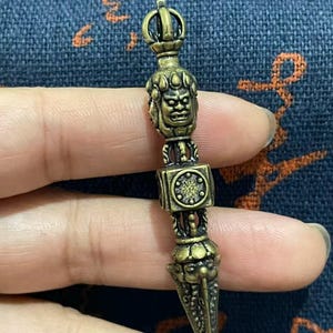 May include: A bronze-colored, ornate pendant with a detailed design. The pendant features a stylized face, a square element with a circular design, and a pointed, dagger-like bottom. The pendant is held in a person's hand.